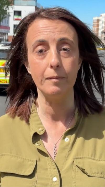 Lucy Manning, a woman with brown hair and a khaki coloured shirt stands with emergency vehicles behind her