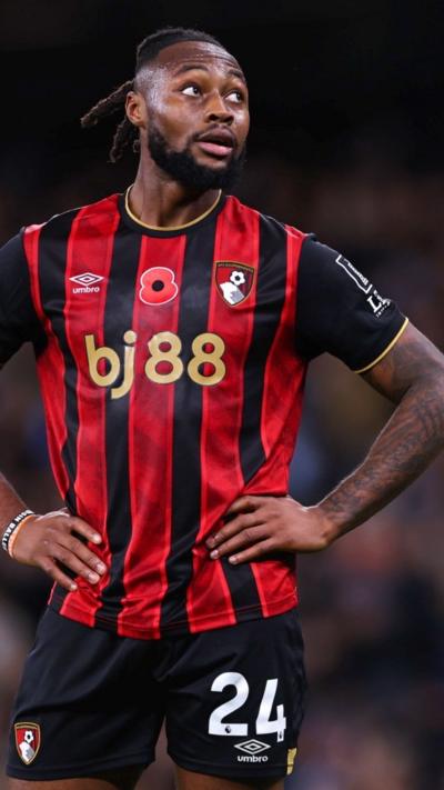 Antoine Semenyo has his hands resting on his hips and looks to the side. He has his hair tied back in a ponytail with dreadlocks. He is wearing an AFC Bournemouth kit with red and black stripes and black shorts which have the number 24 on the leg.