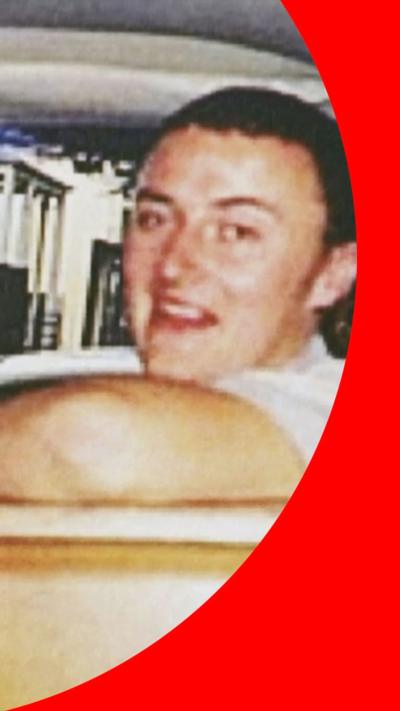 Grainy old photo of Peter Falconio, smiling to camera while leaning against a shelf with his arm up.