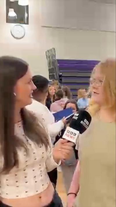 A woman smiles while holding a microphone towards a student with blonde hair and glasses who is also smiling