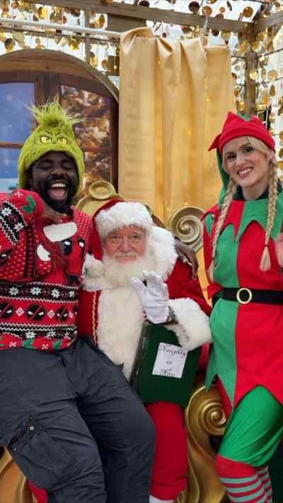 A group of three festive elves dressed in green and red with pointy hats flank Kris Kringle and Kofi Smiles. Kofi is wearing a Christmas jumper and a green Grinch hat and Kris Kringle is an elderly man with a white beard and a red and white suit and a Santa hat. The group are in a grotto with winter scenes and a mock fireplace and sacks of presents.