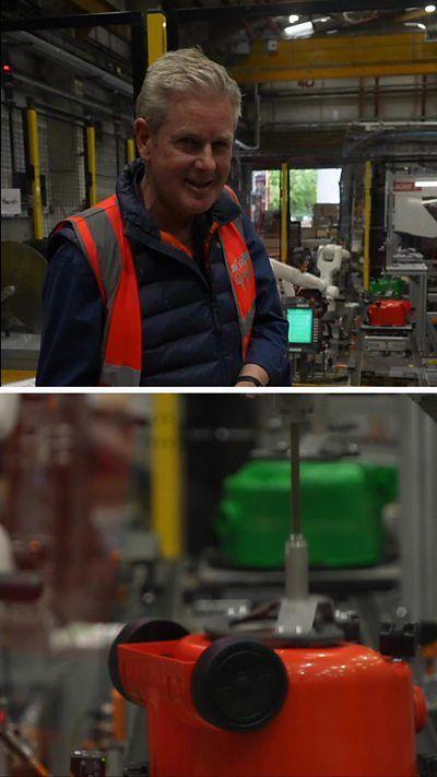 A man stands in a factory which manufactures vacuum cleaners. He is wearing an orange high vis vest on top of a navy blue puffer jacket.