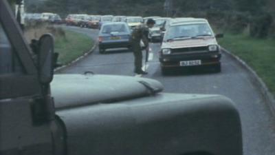 Army officer bends down to speak with driver in a car, and carries out checks along a line of cars that stretches around the road bend.