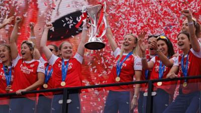 Arsenal footballers hold trophy up. Red and white confetti showers them and they wear medals with their kit