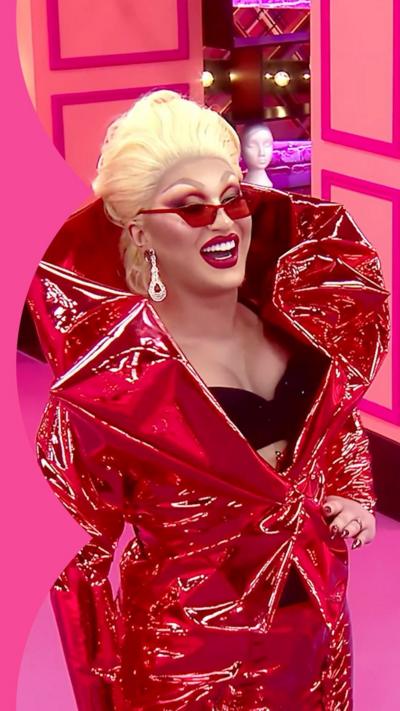 The Vivienne smiles in the Drag Race UK werk room. She wears a red collared outfit, red sunglasses and silver earrings