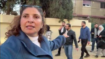 The BBC's Lina Sinjab pointing towards the house belonging to Bashar al-Assad with people walking in front of it