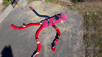 An 18-metre long sock monkey lies on the groun. It has a pink body and red arms and legs. There are people lounging all over the monkey in the sun.