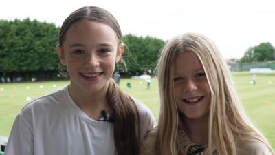 Two girls smiling in front of a cricket pitch background