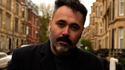 A man with a beard standing in a street with houses behind him.