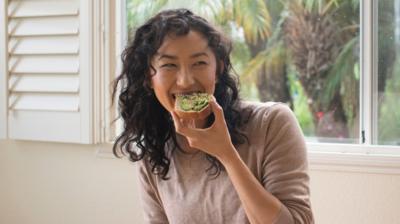 A woman smiles while eating avocado on toast