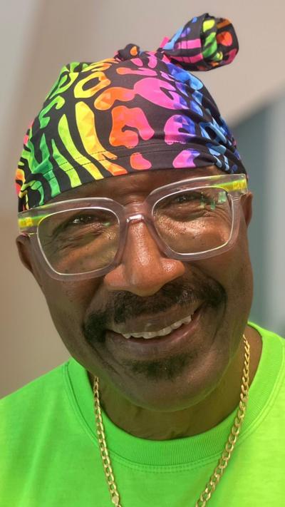 A man wearing a colourful hat and transparent-rimmed glasses and with a moustache smiles towards the camera.