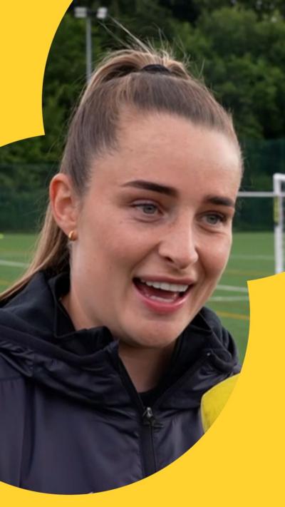 Ella Toone smiles - she has brown hair tied in a ponytail and there is a football pitch behind her 
