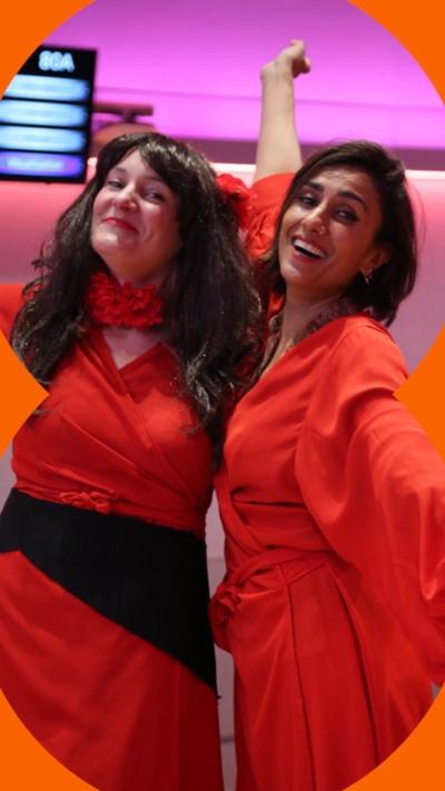 Two women embrace and pose for the camera. They are wearing red dresses like Kate Bush.