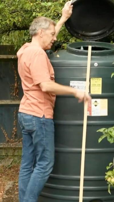 A man with grey hair, orange t-shirt and blue jeans looking inside a green container used to store heating oil