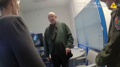 Murdoch wearing dark green jumper stands in front of camera while questioned by two officers in front of him