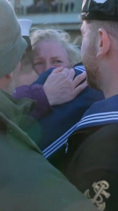 An emotional mother hugging her son, while also looking at her other son. Both men are wearing Royal Navy uniform.