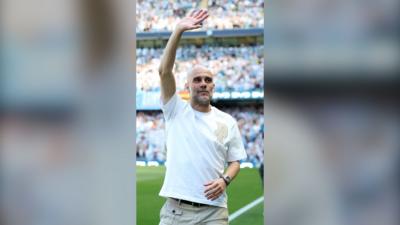 Pep Guardiola in a white shirt with his hand raised in farwell on a football pitch.