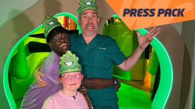 Dr Ronx, Dr Xand and Gracie are stood in front of a pair of giant nostrils which are lit up green. They are all wearing bogey hats. Ronx is hugging Xand and smiling. Xand is holding up one hand and pulling a funny face. Gracie is standing in front of them and smiling.