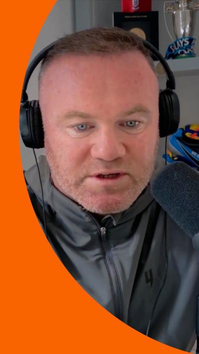 Wayne Rooney has short dark hair and stubble. He is wear a zipped up grey top and headphones.