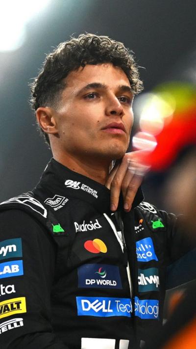 Lando Norris wears a black race suit covered in sponsorship badges and looks away from the camera. He has curly short light brown hair.
