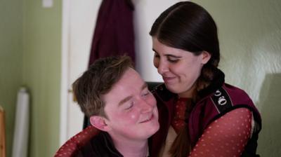 A woman sits on a man's knee, both with coy smiles on their faces