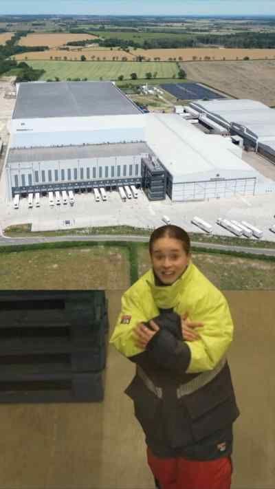 An image of a large warehouse with multiple HGVs outside it taken from an aerial drone. Superimposed is a young woman with cold weather clothing, she is clutching her arms as though shivering. 