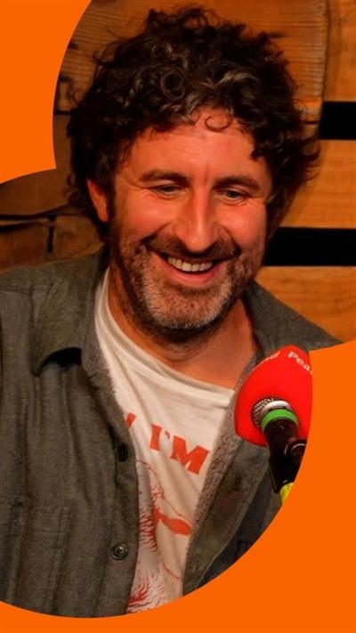 Mark Watson smiles as he talks into an orange microphone.