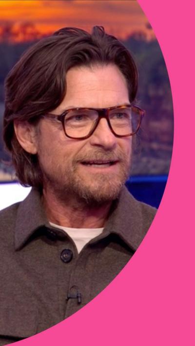 Jason Bateman has long hair swept back and wears glasses and a brown shirt