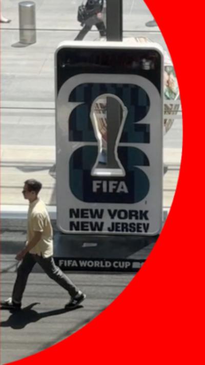 A man walking past what appears to be a face in a whole stand showing the FIFA world cup. It reads 'FIFA New York New Jersey'.