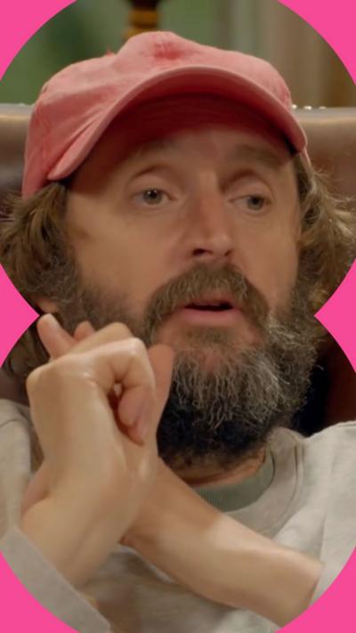 Joe Wilkinson with a very serious expression in a pink cap with his thumb up next to him while he sits in a brown leather chair