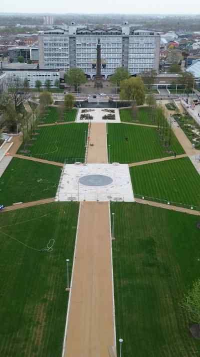 An aerial view of Queen’s Gardens in Hull shows long, symmetrical lawns divided by wide pathways that converge on a central circular plaza, framed by newly planted trees. The gardens are surrounded by a mix of modern and historic buildings and the city stretching out into the distance beyond.