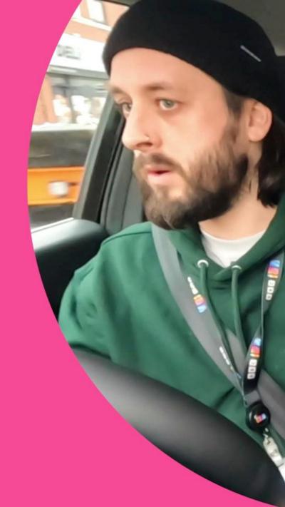 A man with a beard is driving a car, he wears a beanie and a green hooded sweatshirt