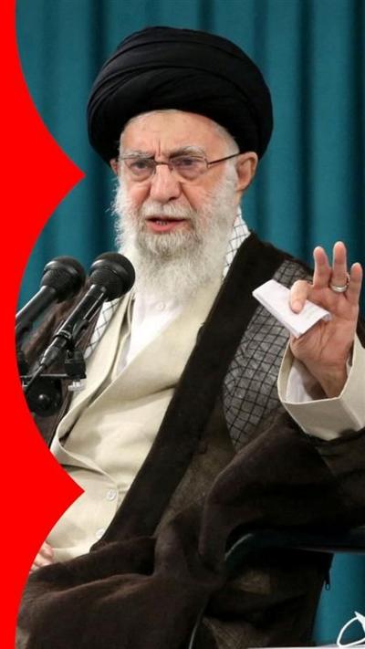 Ayatollah Ali Khamenei in front of microphones with his hand in the air.