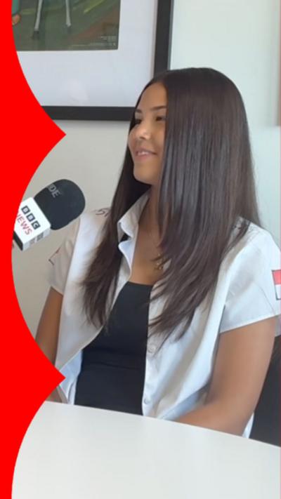 A young girl with brown hair sat in a chair. She's facing a reporter who is holding a mic to her face.