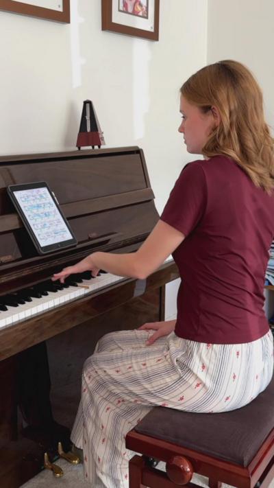 Freya is sitting at a glossy brown piano, with her left hand resting on the keys. Her right hand is resting on her knee, which is where it normally remains while she is performing. She is sitting upright on the piano stool and is wearing a burgundy t-shirt and white trousers with thin blue stripes and a pink dotted pattern.