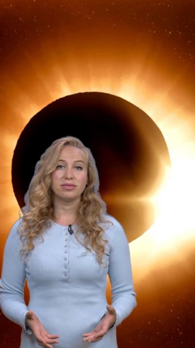 A woman with long blonde hair wearing a blue dress stands in front of a graphic of the Moon moving across the Sun