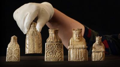 Gloved hand picking up one of five Lewis chessmen. Two are facing the camera and three away from the camera.  