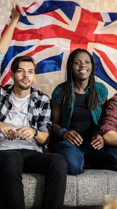 A man with short dark hair, wearing a black and white checked over a white t-shirt, sat next to a woman with long dark hair, wearing a green short-sleeved shirt over a black t-shirt. There is a large Union Jack flag in the background.