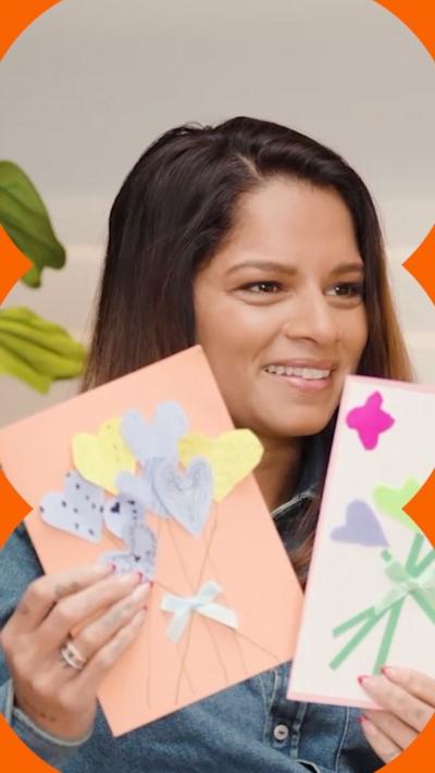 A woman in a jean jacket holds up two greetings cards, made by children.
