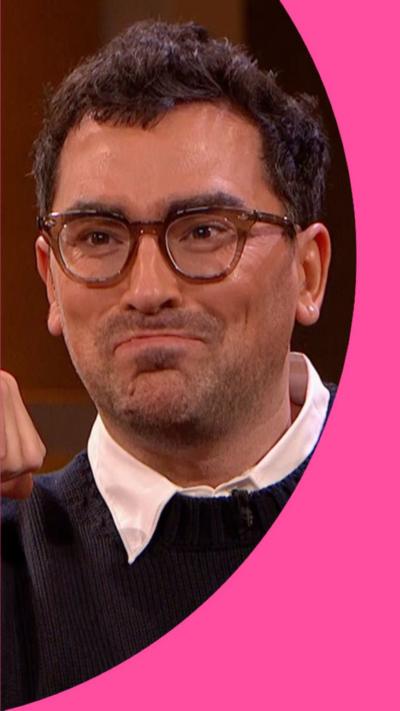 Dan Levy has short dark hair and is wearing glasses and a blue jumper.