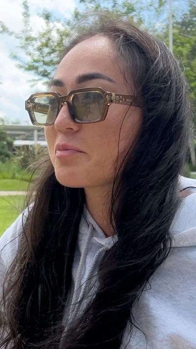 Close-up of a young woman outdoors wearing tinted rectangular sunglasses and a light grey hoodie, with long dark hair falling over the shoulders. Background shows green grass, trees, and a modern structure under a bright daytime sky.