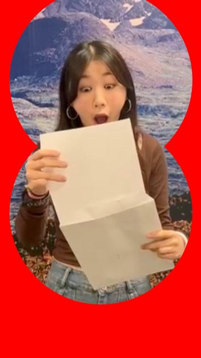 A student looks shocked, reading a piece of paper coming out of an envelope. She has long black hair and wears a brown top and blue jeans