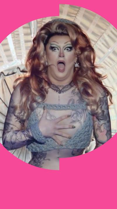 Kate Butch looks shocked with her hand covering her chest. She has long wavy ginger hair and wears a knitted bikini top.