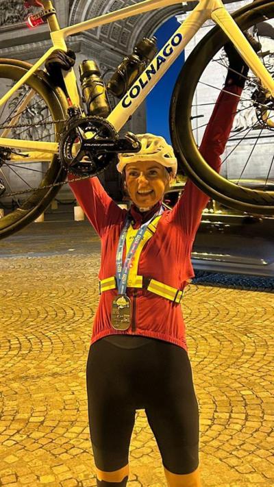 A smiling Lyndsey Blair, raises her bike in triumph after crossing the finish line in Paris. She is wearing black cycle shorts, a red long-sleeved cycle top and a yellow helmet. She has a medal round her neck