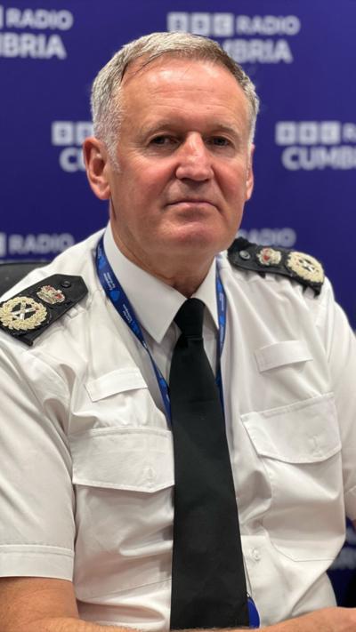 A uniformed police officer wearing a white shirt with black epaulettes, a black tie, and a blue lanyard sits in front of a blue BBC Radio Cumbria backdrop. The officer faces slightly to the side, appearing to be in conversation during an interview.