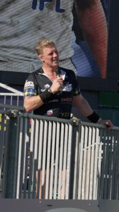 Former England captain Lewis Moody stands behind some railings on a stand at Bath Rugby's Recreation Ground. He's wearing a black cycling outfit with tartan sleeves and black sweatbadnds. He's holding a microphone. There is a big screen in the background showing a photograph of Lewis Moody playing for Bath.