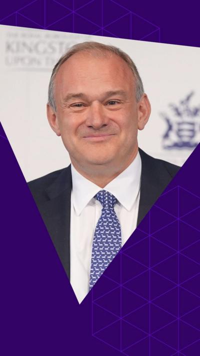 Ed Davey smiles. He wears a dark suit and a blue patterned tie