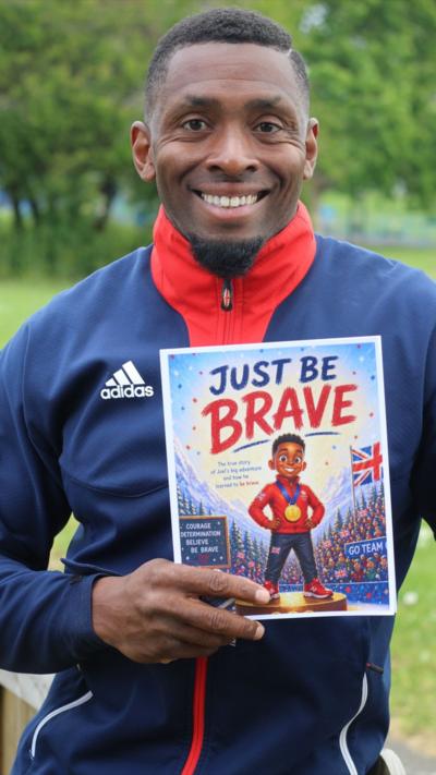 A black man in a blue and red tracksuit top holding a children's book called Just Be Brave