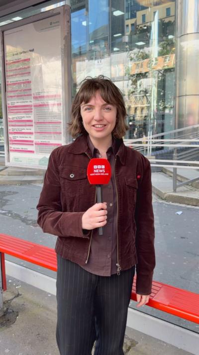 Olivia Copeland has shoulder length brown hair, she is wearing a brown jacket and holding a red microphone at a bus stop. 