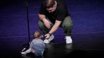 A baby on stage with a man crouching down holding a micophone. The baby is wearing a blue striped top. The man is wearing green trousers with a black tshirt and white trainers.